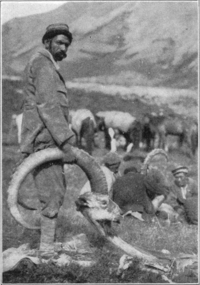 A man holding a horn attached to a disembodied ibex head. Behind are a group of men surrounding another animal with antlers.